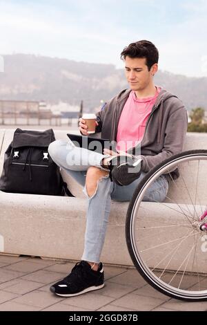 photo verticale d'un jeune homme assis à l'extérieur avec un café à l'aide d'une tablette à côté de son vélo, concept de technologie et de style de vie urbain, espace de copie pour Banque D'Images
