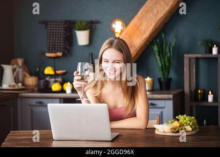 Une jeune femme heureuse boit du vin blanc tout en discutant vidéo avec son petit ami assis dans le salon Banque D'Images