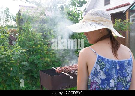 Une fille dans un chapeau de paille prépare de la viande fraîche aromatique sur le gril sur un fond de feuillage vert lors d'une journée d'été lumineuse. Gros plan Banque D'Images