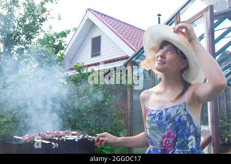Une fille dans un chapeau de paille prépare de la viande fraîche aromatique sur le gril sur un fond de feuillage vert lors d'une journée d'été lumineuse. Gros plan Banque D'Images
