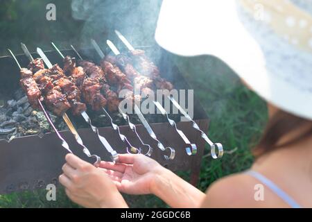 Une fille dans un chapeau de paille prépare de la viande fraîche aromatique sur le gril sur un fond de feuillage vert lors d'une journée d'été lumineuse. Gros plan Banque D'Images