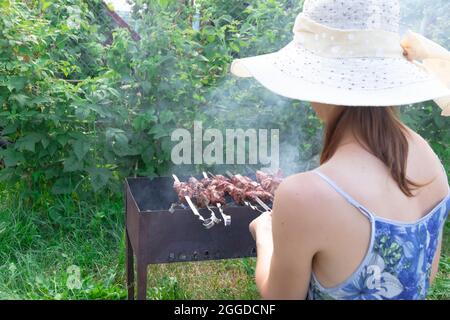 Une fille dans un chapeau de paille prépare de la viande fraîche aromatique sur le gril sur un fond de feuillage vert lors d'une journée d'été lumineuse. Gros plan Banque D'Images