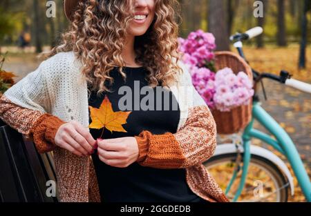 Vue rapprochée d'une femme souriante se reposant dans la nature à côté d'un vélo en stationnement. Le cycliste tenant dans ses mains jaunit la feuille d'érable tombée et se réjouissant de la beauté de l'automne doré. Banque D'Images