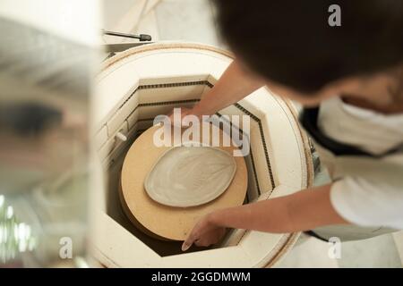 De ci-dessus, crop anonyme femme artisan plateau de mise avec plaque d'argile non cuite dans four spécial de four tout en travaillant dans le studio de poterie Banque D'Images