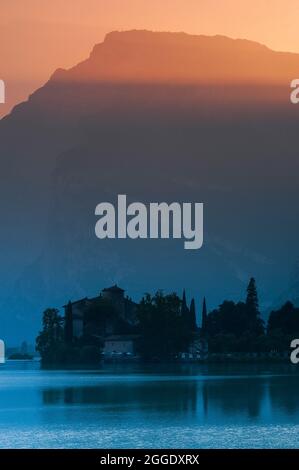 Une soirée calme de septembre dans les Dolomites. Les rayons horizontaux du soleil couchant baignent les hauteurs accidentées de Monte Casale dans une lumière orange-rose douce, contrastant avec des ombres et des reflets bleu doux sur la surface d'un lac, le Lago di Toblino, dans le Trentin-Haut-Adige, en Italie. À mesure que la lumière du jour s'estompe, le détail du château médiéval de Toblino, ancienne résidence d'été des prince-évêques de Trent, avec son contour se tournant vers la silhouette. Banque D'Images