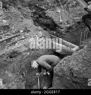 Un homme qui creuse dans la tranchée de la calotte de la pile sur le chantier de construction de la cathédrale de Coventry, avec un cercueil de pierre derrière lui qui dépasse dans la tranchée. Cette photographie a été prise au début de la construction de la cathédrale de Coventry, qui comprenait la démolition, le dégagement du site, l'excavation et la conduite de pieux avec les premières fondations posées le lundi 7 mars 1955. Banque D'Images