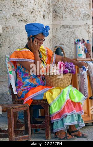 Femme, cubaine, en costume traditionnel multicolore, assise sur une chaise et parlant sur un téléphone portable, Vieille ville, la capitale de la Havane, la Havane Banque D'Images