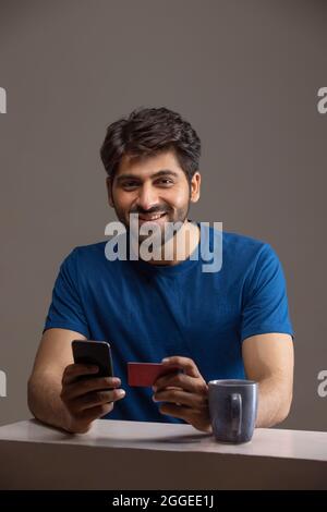 PORTRAIT D'UN JEUNE HOMME QUI REGARDE AVEC JOIE L'APPAREIL PHOTO TOUT EN FAISANT UNE TRANSACTION EN LIGNE Banque D'Images