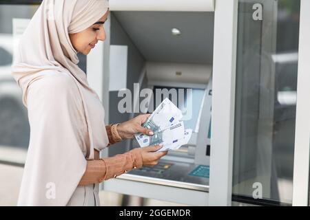 Bonne jolie femme islamique millénaire dans le hijab comptant beaucoup d'euros près de atm extérieur. Transfert de fonds, compte financier personnel, réception d'argent, W Banque D'Images