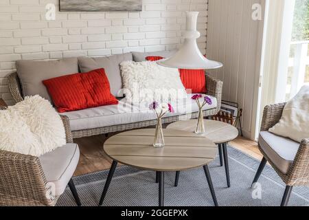Canapé en rotin avec deux fauteuils et oreillers rouges et blancs confortables, mur en pierre blanche derrière, style scandinave. Banque D'Images