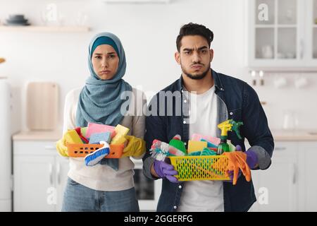 Lassé mari et femme de famille musulmane nettoyant leur nouvel appartement, tenant des fournitures de maison, intérieur blanc de cuisine, espace de copie. Entretien de la maison s Banque D'Images