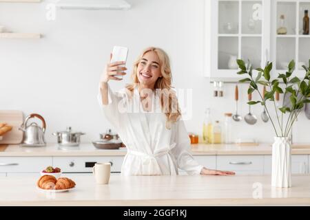 Selfie du matin, blog vidéo et appel, distance sociale. Souriant belle femme européenne millénaire en peignoir de boire du café et de prendre selfie sur sma Banque D'Images