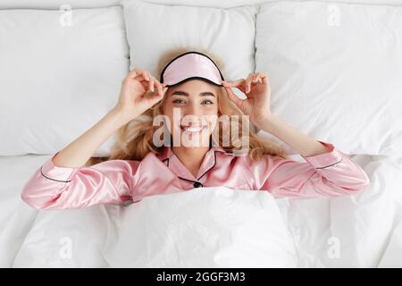 Il est temps d'aller au travail, de se réveiller tôt le matin, de se reposer et de se détendre à la maison. Heureuse belle jeune femme européenne en pyjama rose prend son masque de sommeil, l Banque D'Images