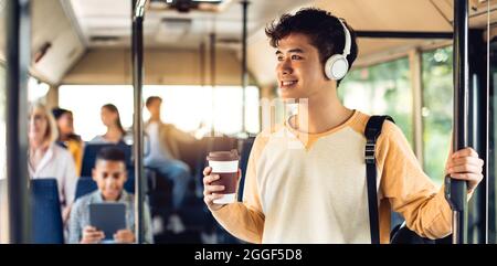 Style de vie des jeunes. Un homme asiatique positif se tenant dans les transports publics, portant un casque tenant une tasse à emporter et une poignée, buvant du café, conduisant à u Banque D'Images