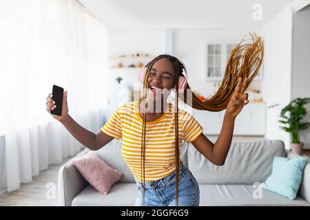 Émotive jeune femme noire amante de musique appréciant le tout nouveau casque stéréo, danse et gestuelle tout en écoutant de la musique, en utilisant l'application mobile Banque D'Images