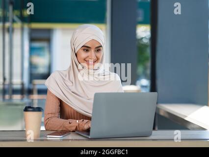 Les nomades numériques travaillent et étudient à distance, des dispositifs pour la pigiste en extérieur. Joyeuse millénaire belle dame arabe dans hijab assis à table, dactylographiant sur le lapte Banque D'Images