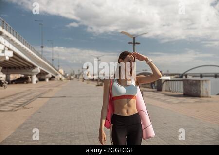 Belle dame dans des vêtements de sport élégants avec tapis roulé regarde dans la distance sur le remblai de ville Banque D'Images