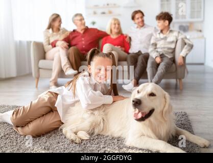 Petite fille mignonne jouant avec le chien Golden Retriever sur le sol dans le salon, ses parents se reposant sur le canapé à la maison. Grande famille multi-génération spendi Banque D'Images