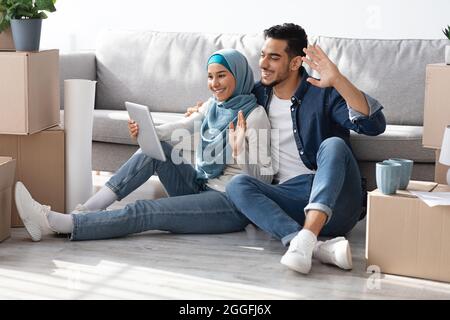 Heureusement marié jeune couple arabe ayant la fête en ligne avec des parents, mari et femme dans hijab montrant leur nouvel appartement, assis au sol parmi Banque D'Images