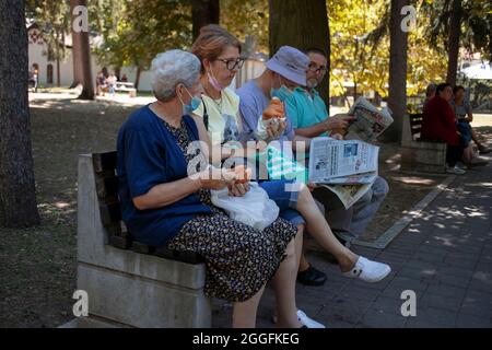 Sokobanja, Serbie, 19 août 2021: Deux femmes assises sur un banc ayant un snack à côté des hommes lisant le journal Banque D'Images