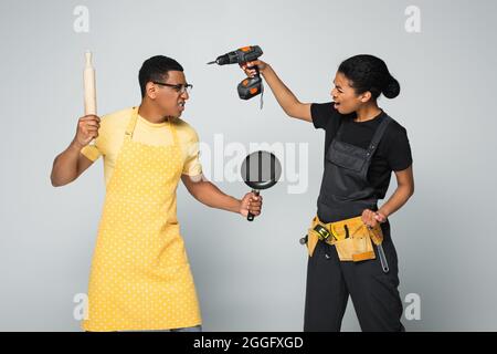 homme américain africain en colère dans un tablier tenant une goupille et une poêle à frire tout en luttant avec une femme de réparation isolée sur gris Banque D'Images