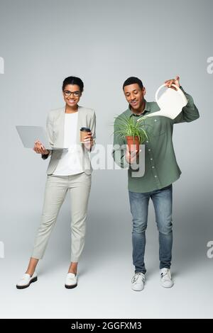 bonne femme d'affaires afro-américaine tenant un café à emporter et un ordinateur portable près d'un arrosoir de jardinier sur fond gris Banque D'Images