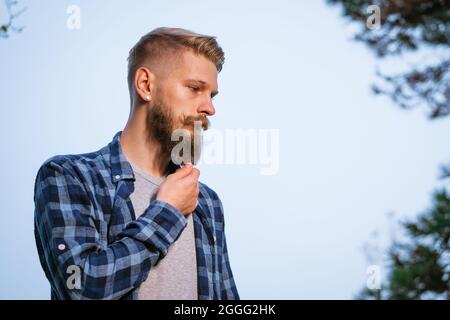 Un jeune homme caucasien barbu dans une chemise à carreaux voyage seul dans la forêt d'automne et regarde sur le côté. Randonnée, voyage en plein air, loisirs et aventure active concept Banque D'Images