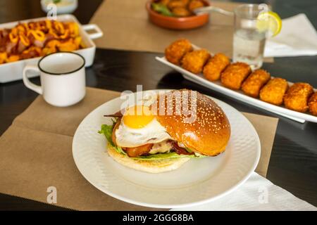 Hamburger avec œuf frit, bacon frit et fromage fondu servi dans un restaurant de tapas avec d'autres plats tels que croquettes et chorizos à l'enfer Banque D'Images