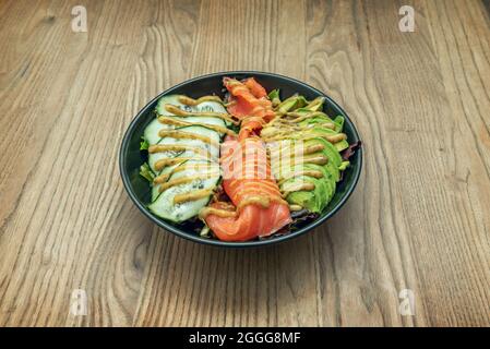 Spectaculaire bol à poke noir avec tranches de saumon fumé, tranches de concombre, avocat à moitié roulé, pousses de laitue et sauce avec aneth sur une table en chêne Banque D'Images