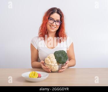 La femme caucasienne préfère une alimentation saine. REDHEAD fille choisit entre brocoli et beignes sur fond blanc. Banque D'Images