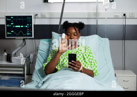 Malade afro-américain utilisant la technologie d'appel vidéo sur le smartphone se rétablissant dans le lit de l'hôpital. Jeune personne atteinte de maladie agitant tout en étant sur Internet conférence en ligne avec la famille Banque D'Images