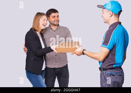 photo de trois personnes, messager en uniforme bleu et montre intelligente a livré un colis à un beau jeune couple heureux. Banque D'Images