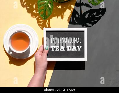 Le texte de la craie blanche Intrnational Tea Day est en main sur le tableau noir. Tasse de thé et plante monstère avec de fortes ombres longues. Flat lay, vue de dessus sur la découpe Banque D'Images