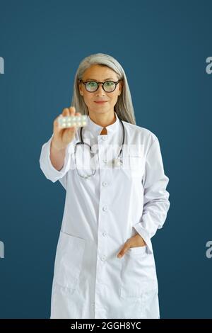 Gaie femme mature médecin en robe blanche montre des comprimés blister posant sur fond bleu Banque D'Images