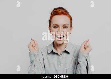 Heureuse femme aux cheveux rouges excitée tenant des poings serrés et célébrant le succès en studio Banque D'Images