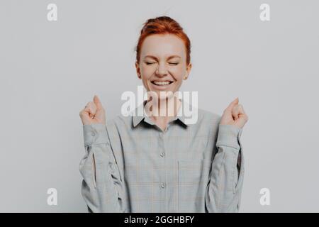 Excitée et surjoyeuse jeune femme au gingembre levant les mains avec des poings serrés, en gardant les yeux fermés Banque D'Images