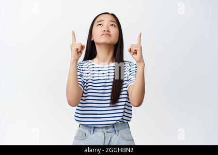 Jeune fille asiatique triste exprime regret et déception, pointant les doigts vers le haut et regardant bouleversé, se tenant contre fond blanc Banque D'Images