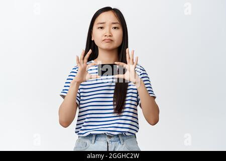 Image de la fille asiatique de brunette frontons les sourcils et les bouts déçus, montre la mauvaise carte de crédit, pas d'argent, debout sur fond blanc Banque D'Images