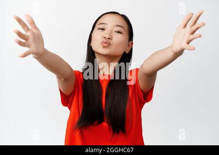 Portrait d'une jolie fille asiatique embrassant, atteignant le câlin, étirant les mains pour embrasser, se tenant sur fond blanc dans un t-shirt rouge Banque D'Images