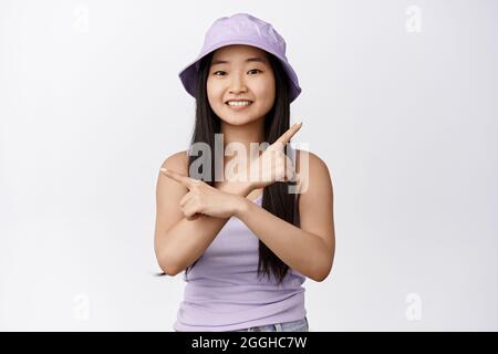 Portrait de mignonne asiatique sourires et points sur les côtés, gauche et droite, montre deux options, se tient dans le chapeau de seau et le débardeur sur fond blanc Banque D'Images