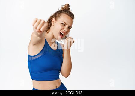 Une fille de forme physique motivée punit l'air, crie et fait des mouvements de boxe, combat dans sportsbra, séance d'entraînement dans la salle de gym, debout sur fond blanc Banque D'Images