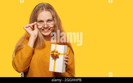 Souriante belle blonde jeune femme portant des lunettes tenant une boîte cadeau isolée sur fond jaune. Joyeux anniversaire, Noël, Saint Valentin Banque D'Images