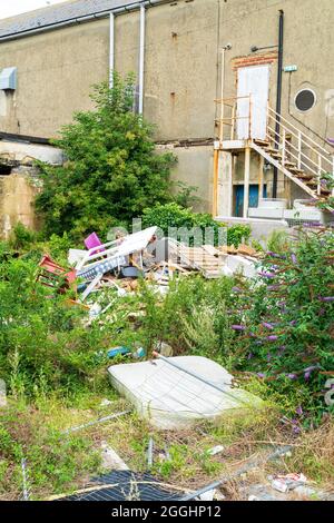 Basculement en vol. Des meubles et des objets de maison à bout volent illégalement sont jetés dans une cour surcultivée déserte d'un bâtiment d'affaires fermé. Banque D'Images