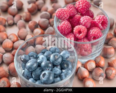 Gros plan de framboises mûres fraîches et de bleuets dans des verres peu transparents et de noisettes fermées sur fond flou. Banque D'Images