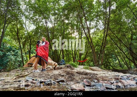 Faible angle d'homme et de femme gaies qui s'embrasse tout en se tenant près de la tente avec le chien en forêt Banque D'Images