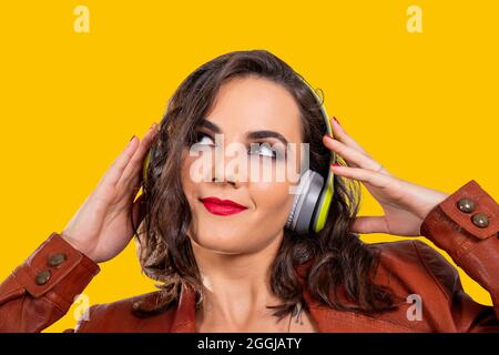 Portrait en studio d'une jeune femme attirante dans une veste en cuir marron écoutant de la musique avec son casque sur fond jaune vif. B Banque D'Images