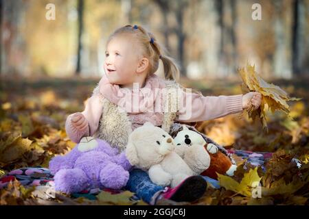 Petite belle fille avec boucles d'oreilles et feuilles d'érable jaune d'automne à la main lapin ensoleillé sur le visage dans le parc d'automne sur les feuilles dorées avec des jouets doux Banque D'Images