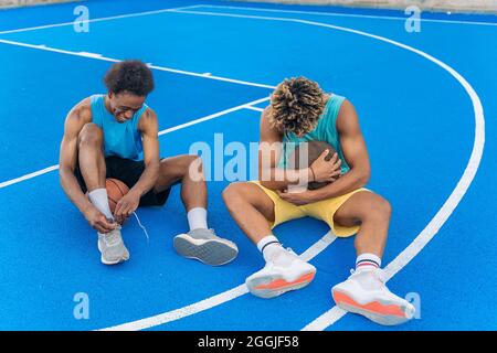 Deux jeunes amis afro-américains se détendent sur le terrain de basket-ball de rue après le match. L'un d'eux nouant ses lacets et l'autre embrassant la balle. Banque D'Images