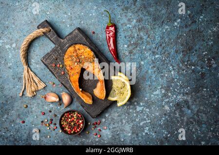 Vue de dessus du steak de saumon au poisson rouge grillé sur un panneau en bois noir avec épices, ail, citron, mélanger le poivre dans un bol sur fond de béton bleu Banque D'Images