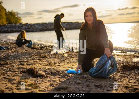 Une jeune femme concentrée qui nettoie la plage avec une équipe de bénévoles pendant le coucher du soleil Banque D'Images